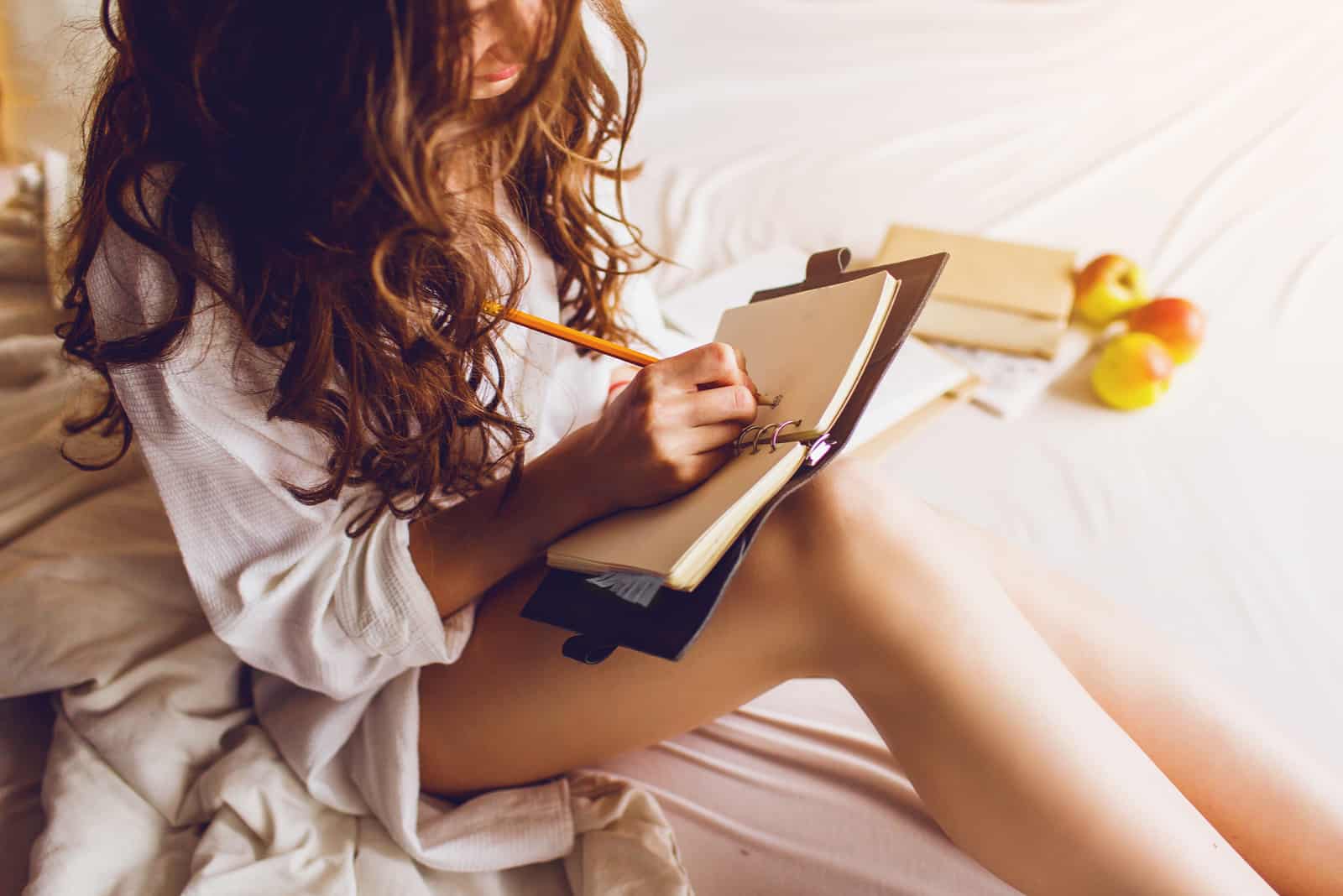 une femme aux longs cheveux bruns est assise dans son lit et écrit