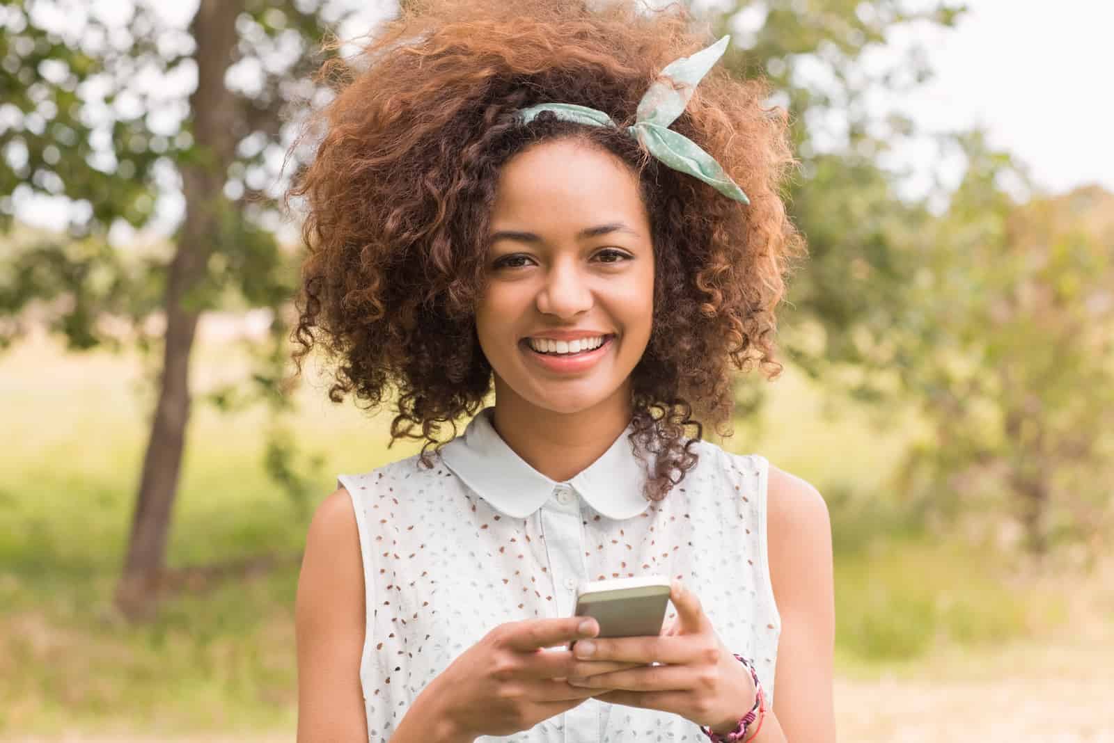 une femme souriante aux cheveux crépus se tient avec un téléphone à la main