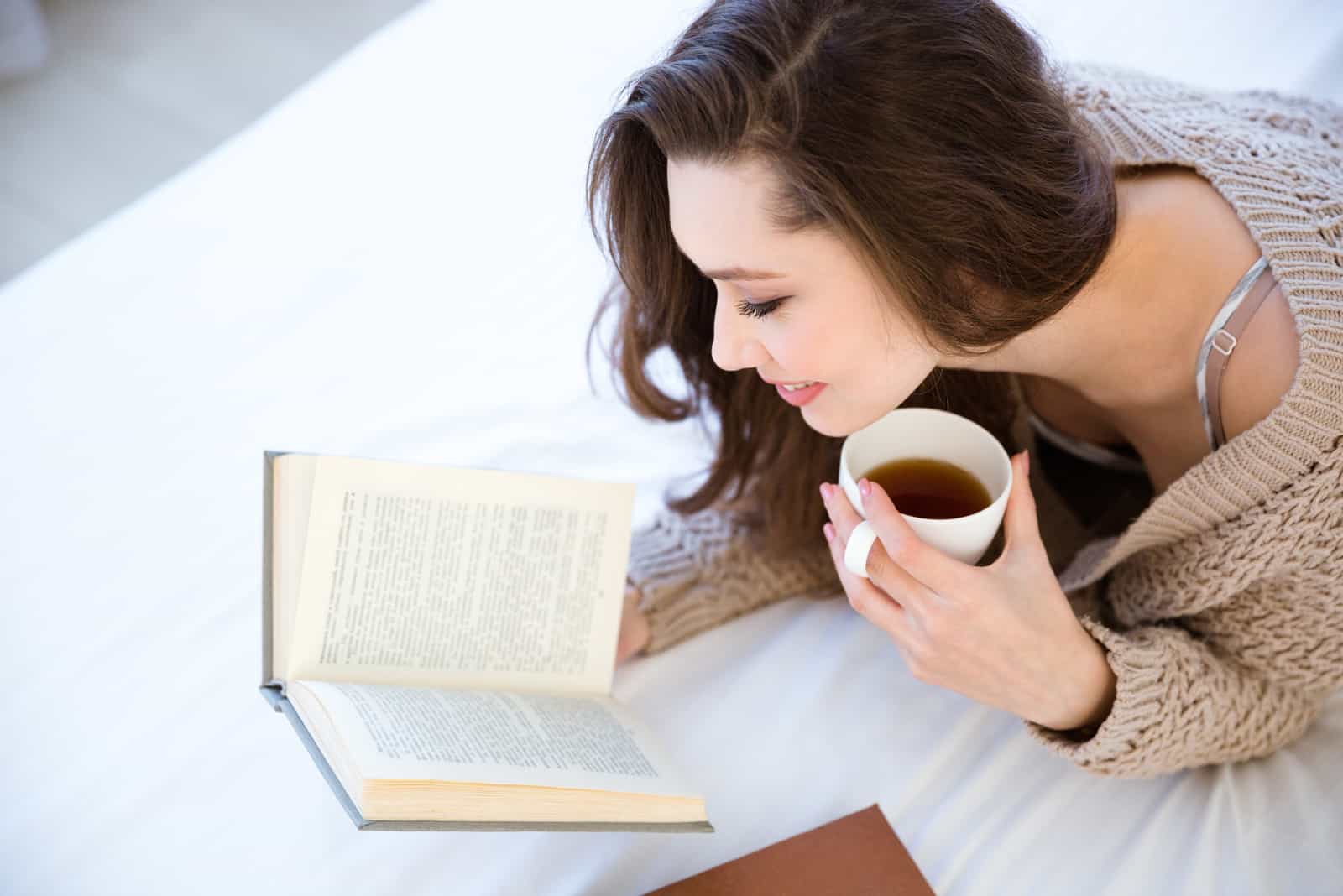 une femme souriante se couche en buvant du café et en lisant un livre