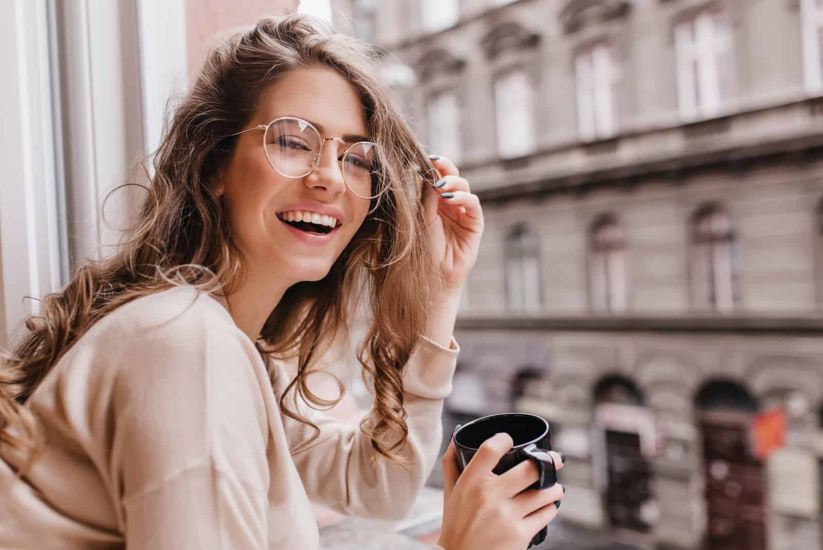 une femme souriante avec des lunettes boit du café