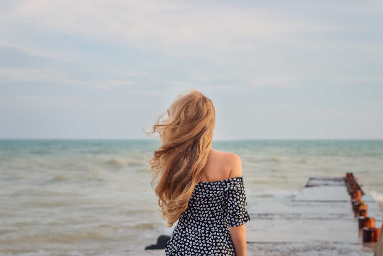 une femme aux longs cheveux blonds se tient le dos tourné et regarde vers la mer