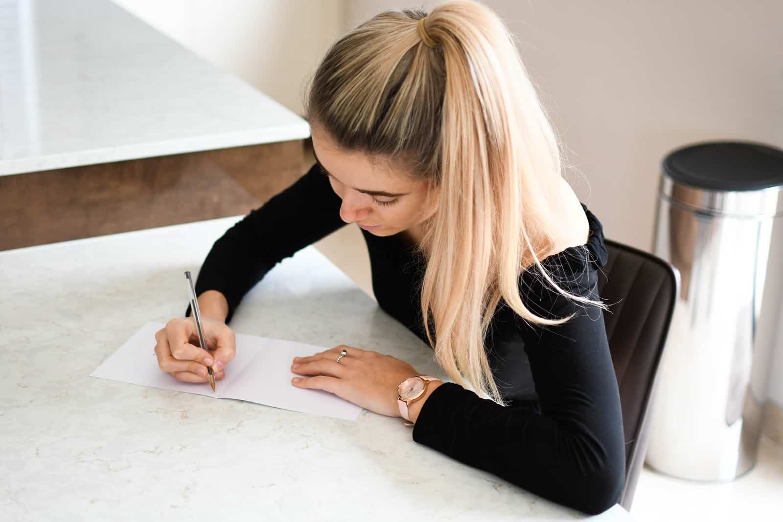 une femme aux cheveux blonds attachés est assise à un bureau et écrit