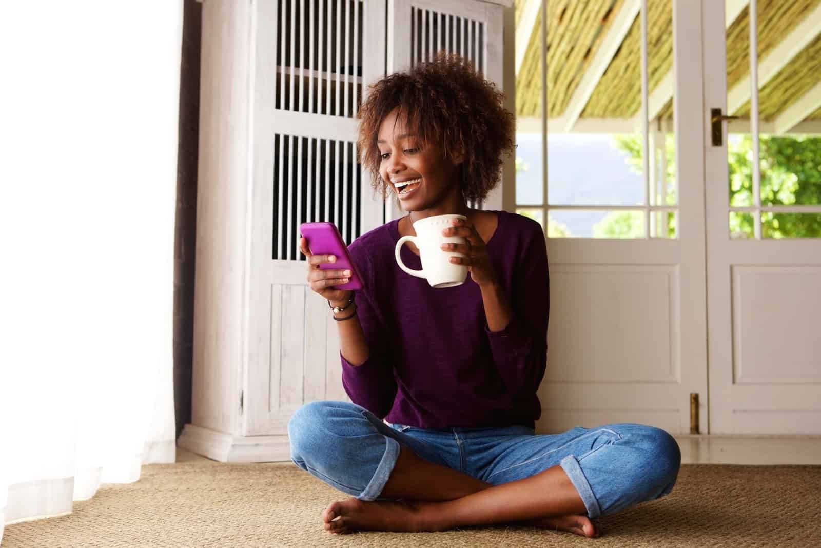 une femme souriante assise par terre buvant du café et tapant au téléphone