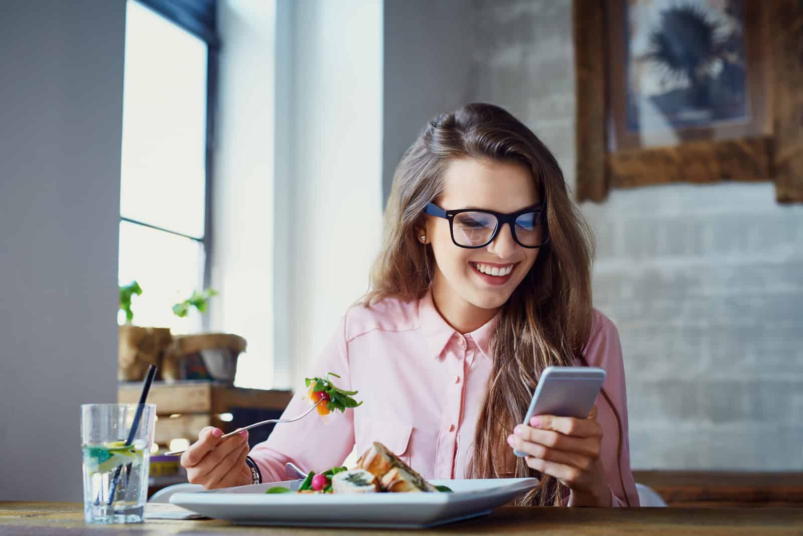 une femme souriante aux longs cheveux bruns est assise dans un café en train de manger et de taper au téléphone