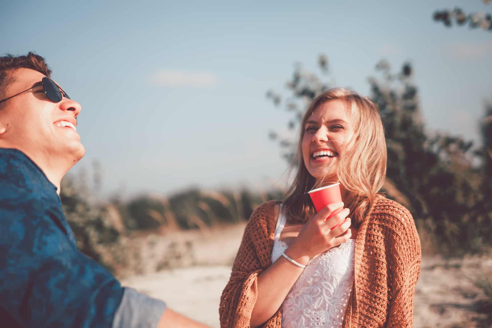 une femme souriante est assise sur la plage et parle à un homme en buvant du café