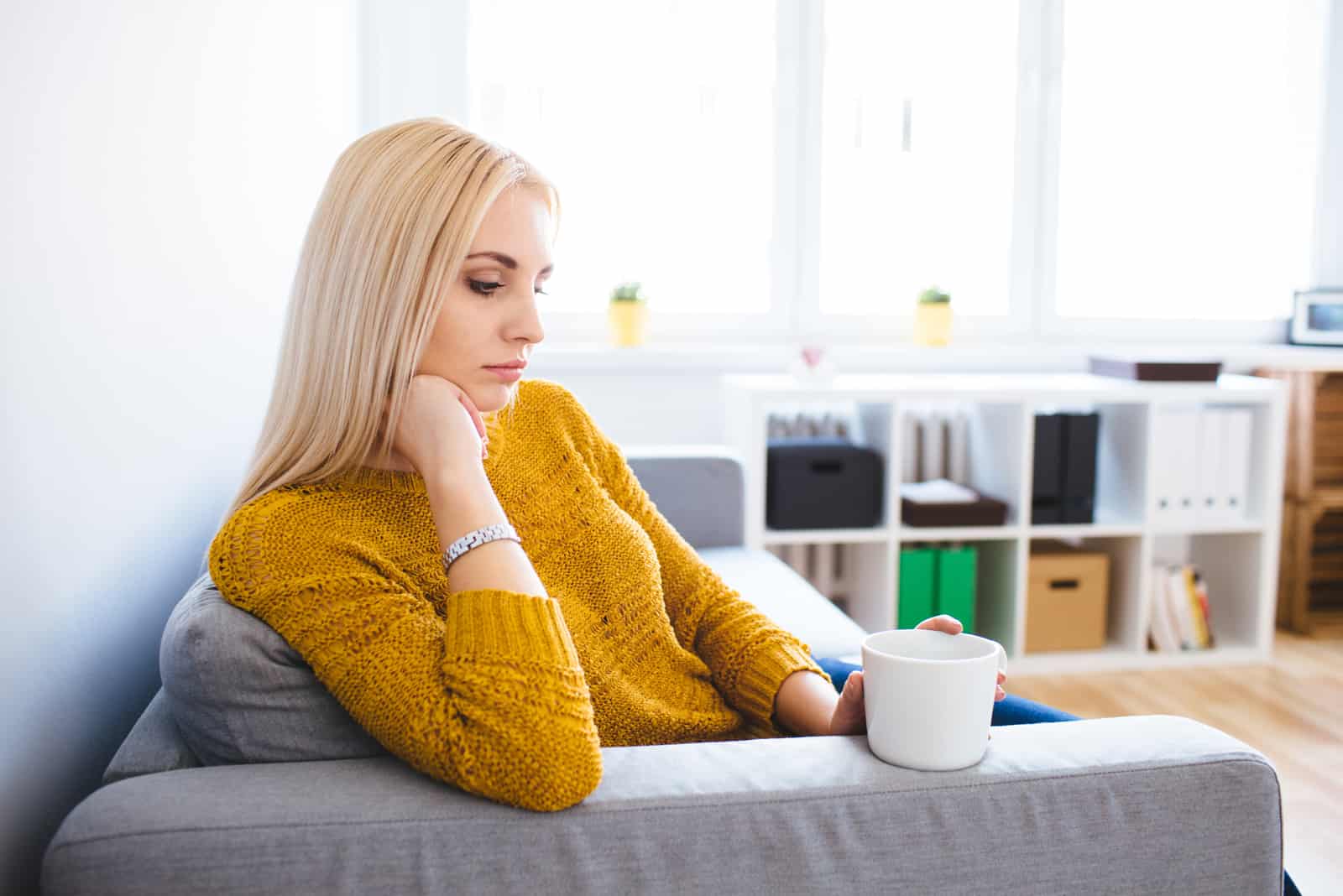 une triste femme aux cheveux blonds est assise sur le canapé et boit du café