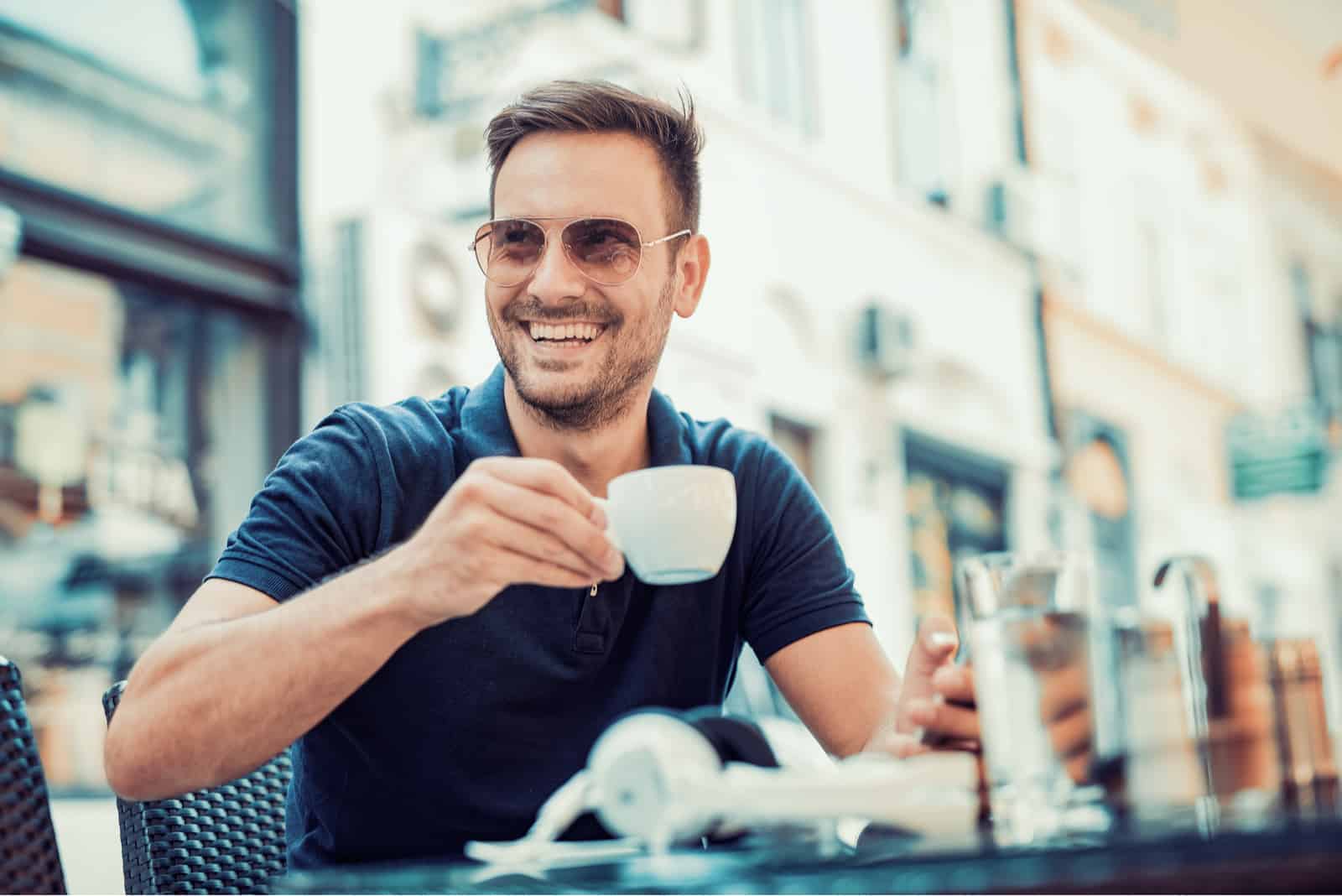 un homme souriant avec des lunettes sur la tête est assis à l'air libre tenant une tasse et regarde ailleurs