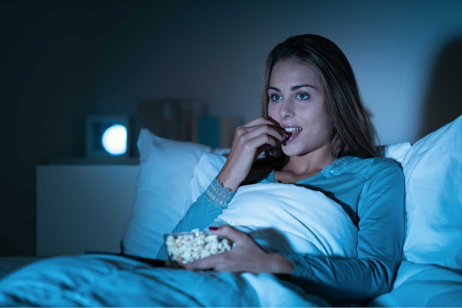 une femme aux longs cheveux bruns est allongée dans son lit en train de manger du pop-corn et de regarder la télévision