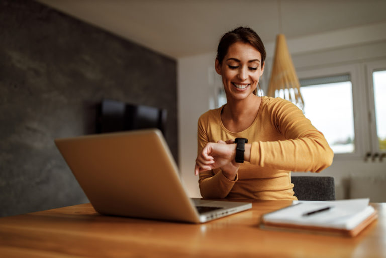 une femme souriante aux cheveux attachés est assise à la table et regarde sa montre