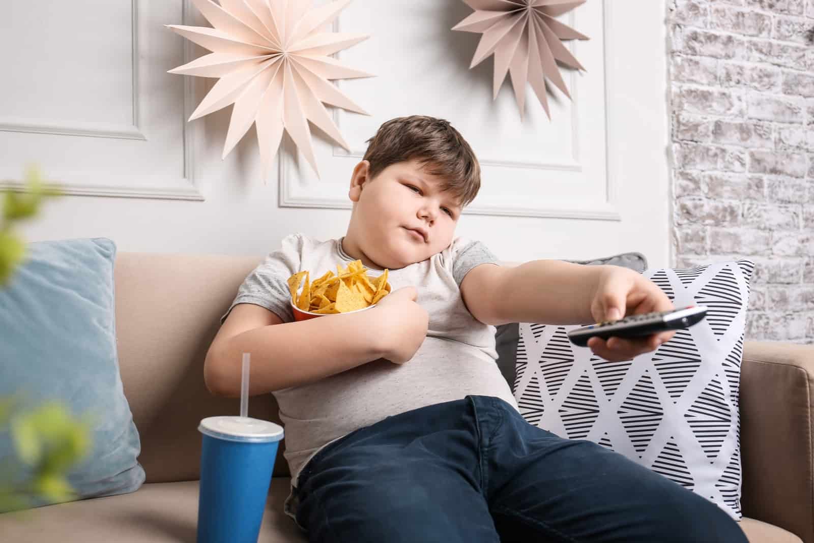 un gros homme est assis sur le canapé en train de regarder la télévision et de manger des chips