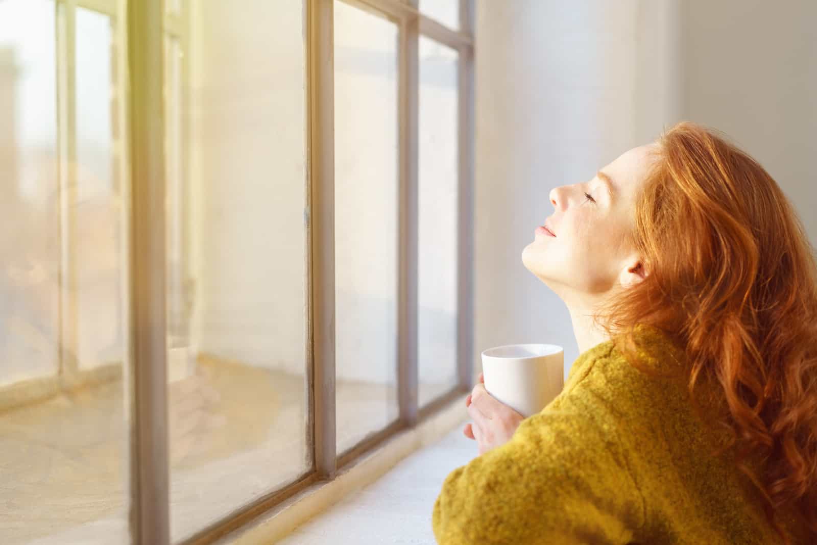 une femme assise près de la fenêtre tenant une tasse à la main et regardant le ciel