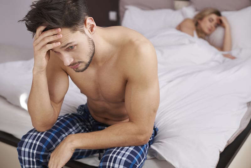 un homme réfléchi assis sur le bord du lit pendant que sa petite amie dort dans le lit