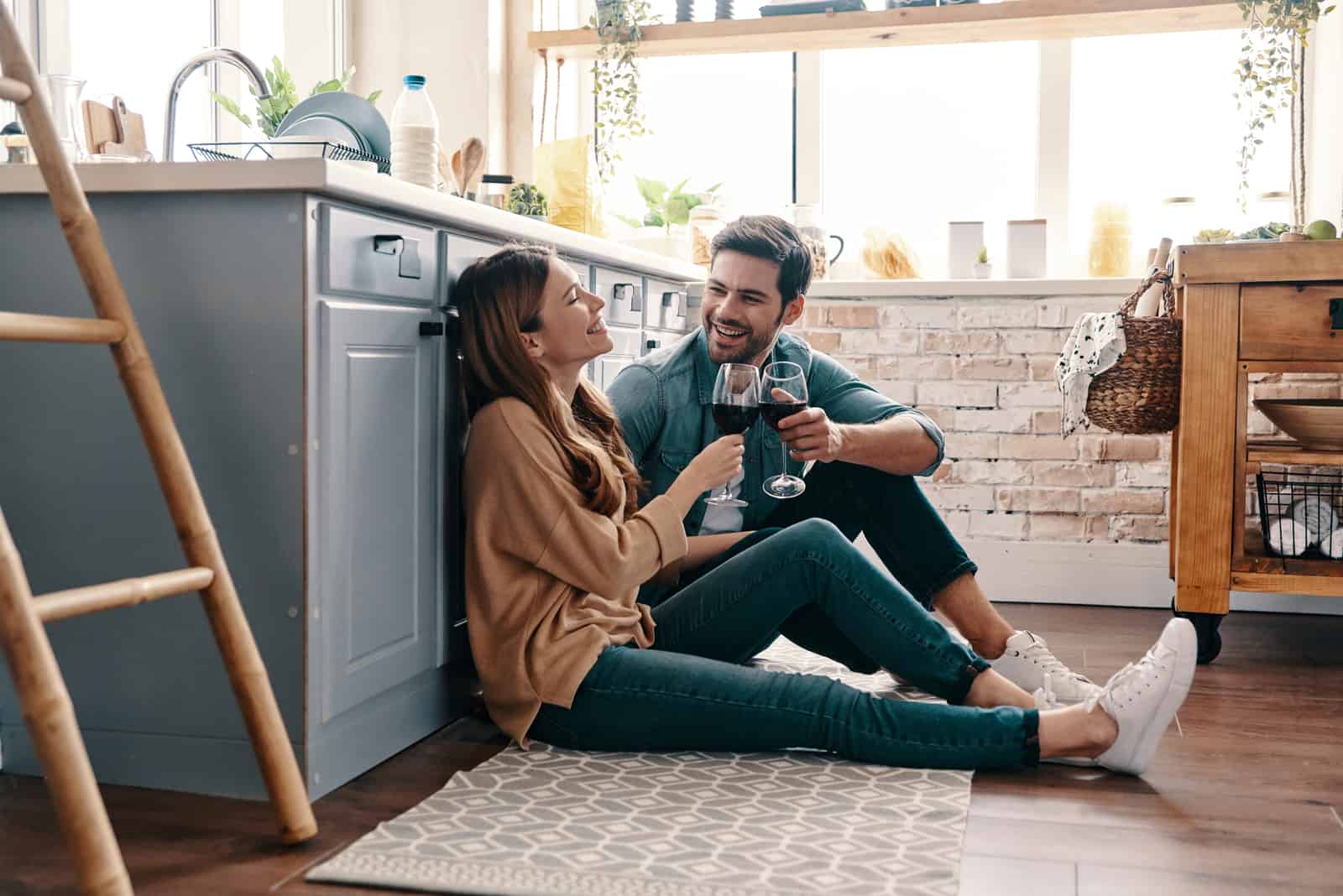 un homme et une femme assis sur le sol de la cuisine buvant du vin et parlant