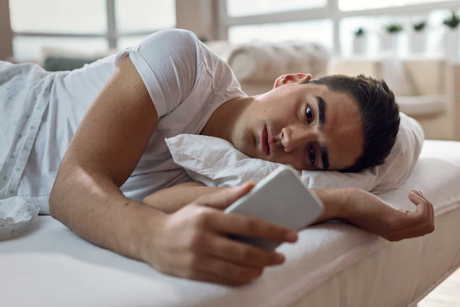 un homme triste couché dans son lit avec un téléphone à la main