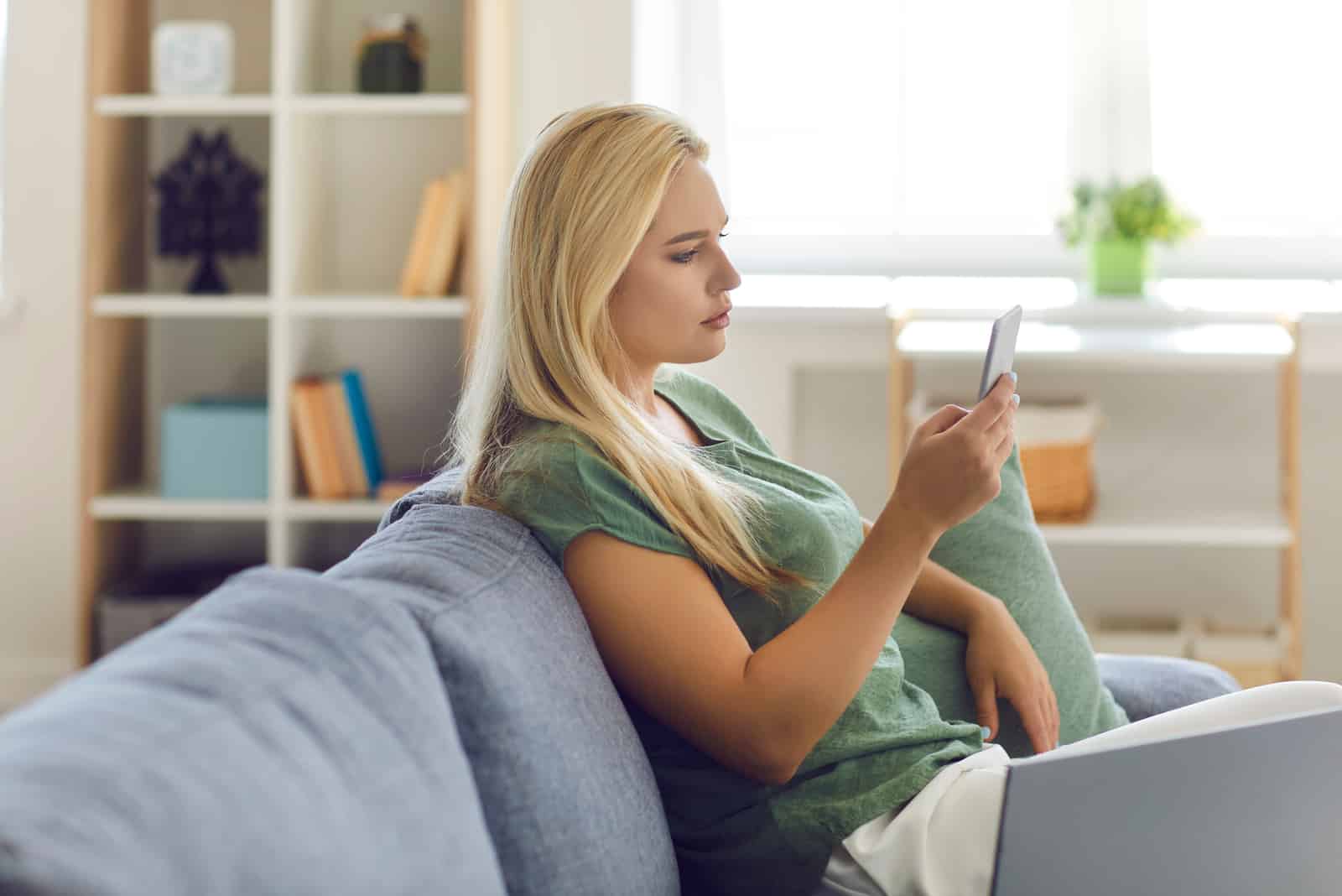 une femme aux longs cheveux blonds est assise sur le canapé tenant un téléphone portable à la main
