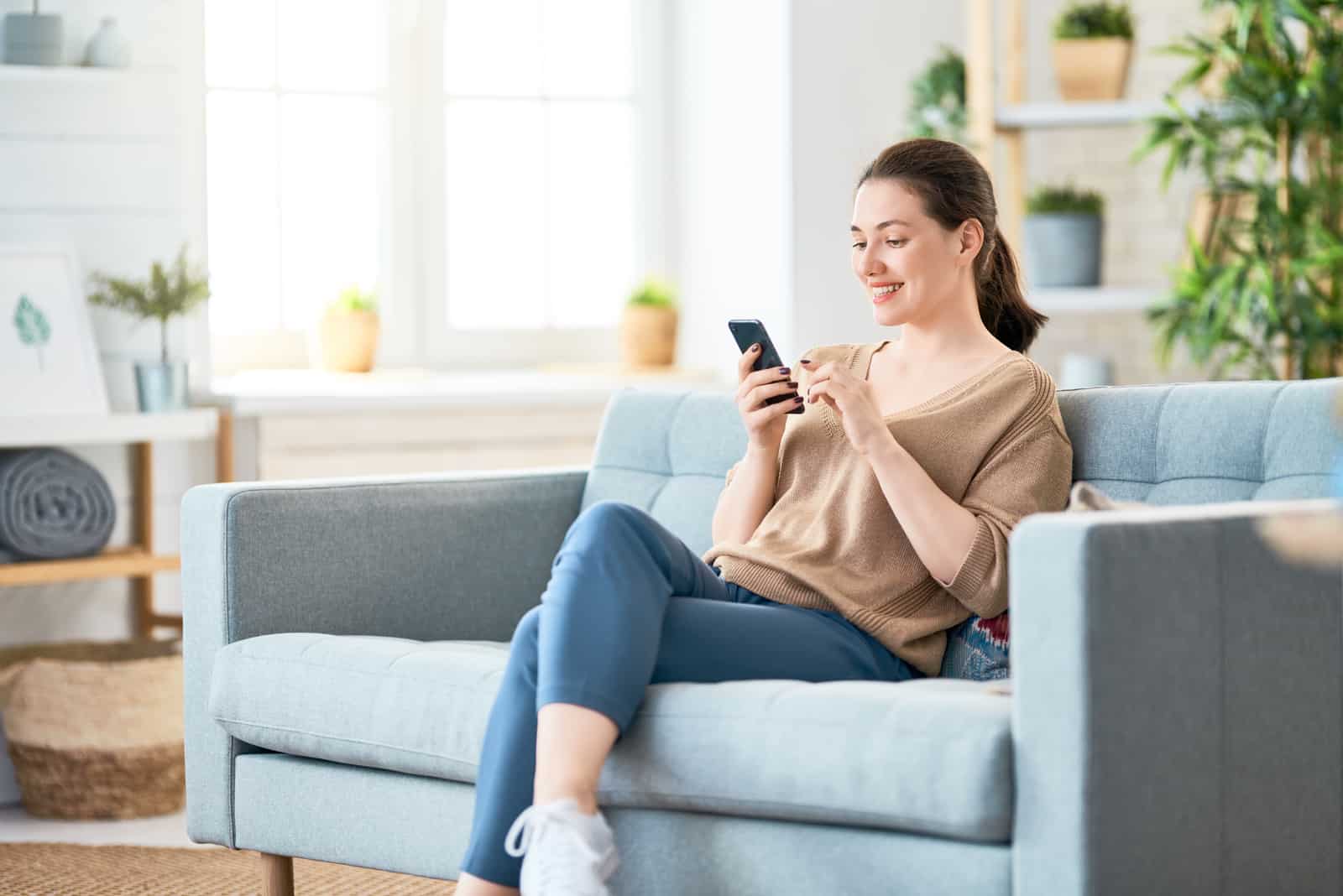 une femme souriante est assise sur le canapé tenant un téléphone à la main