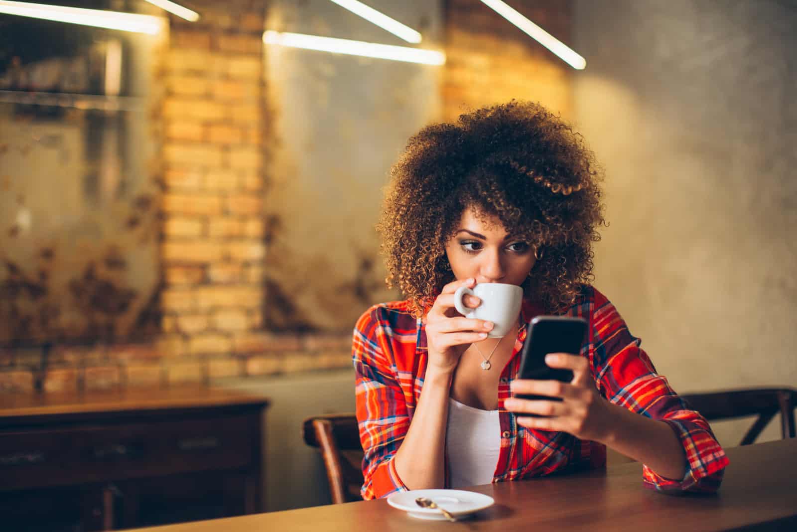 jeune femme aux cheveux bouclés tenant un téléphone et buvant du café