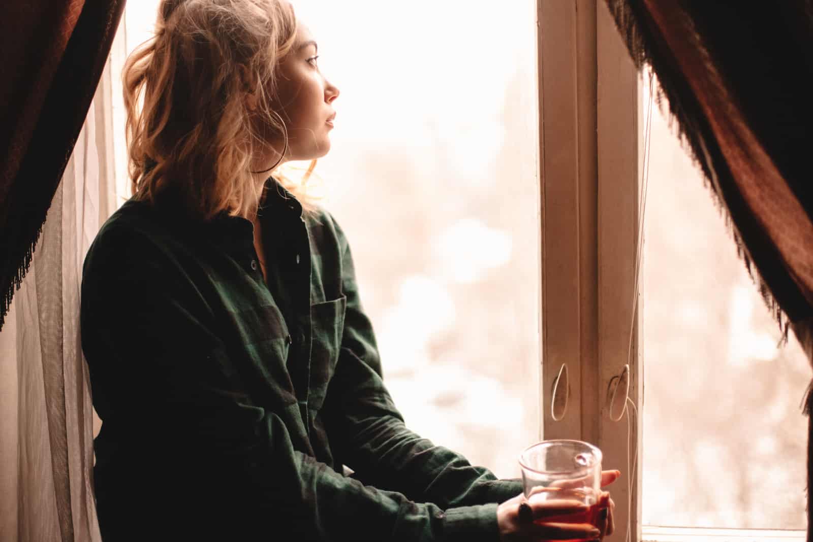 femme blonde assise près de la fenêtre tenant une tasse de thé