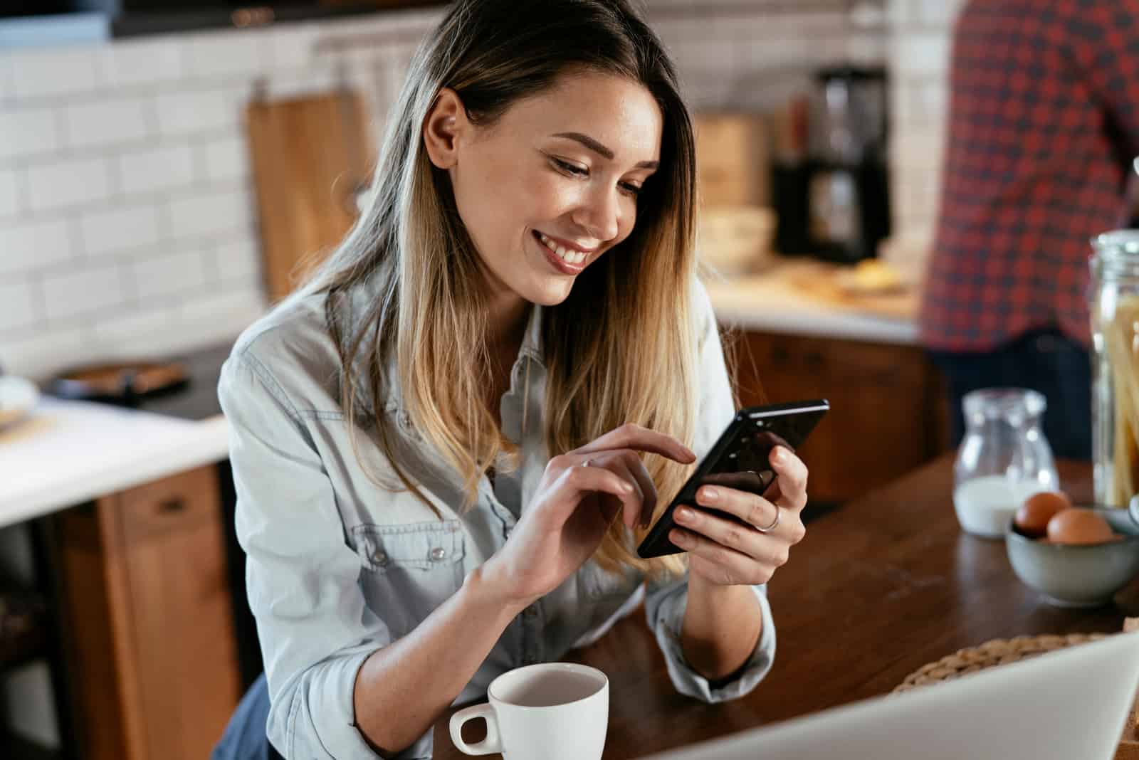 une femme souriante aux longs cheveux bruns est assise à une table tenant un téléphone portable à la main