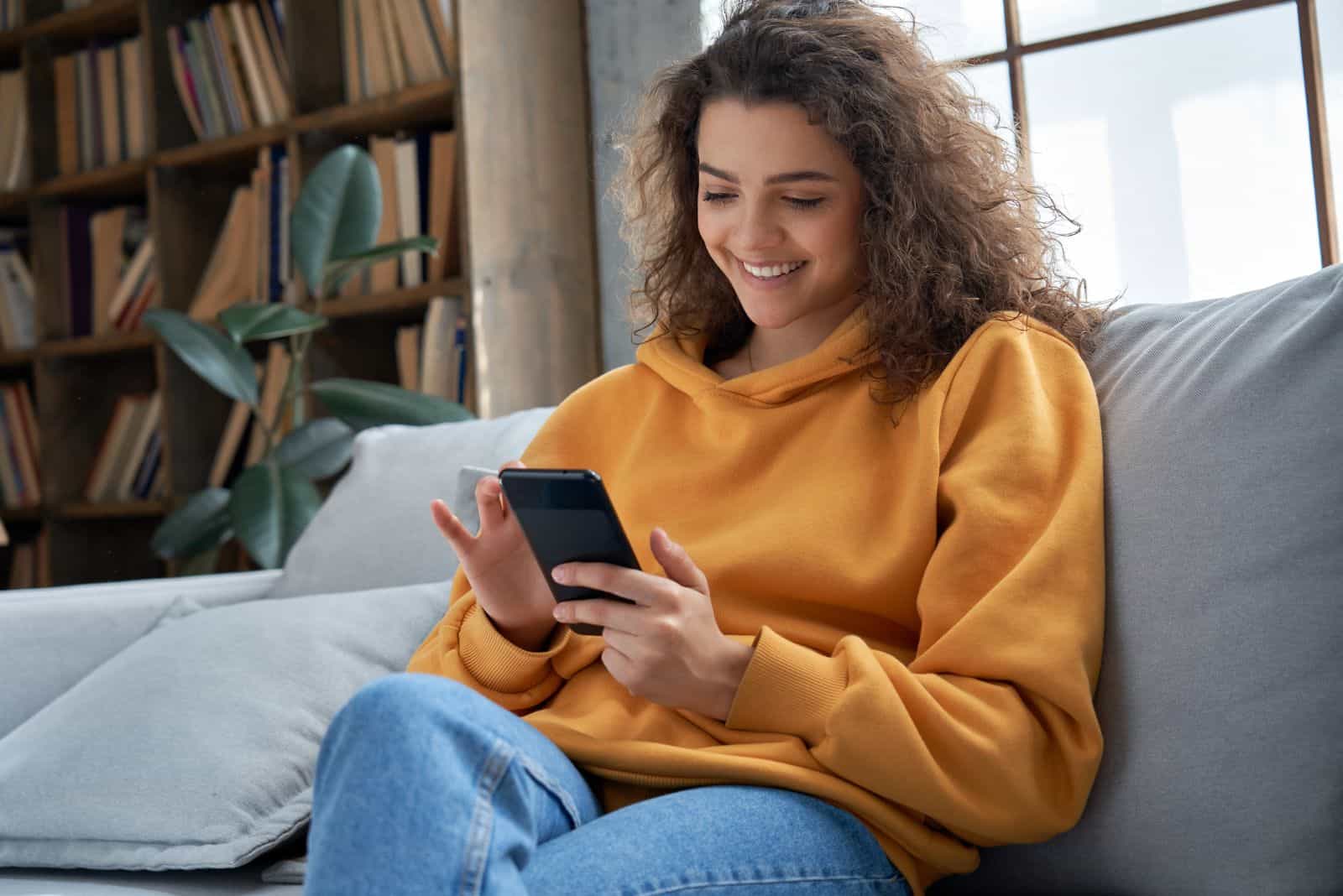 une femme souriante est assise sur le canapé et tape au téléphone