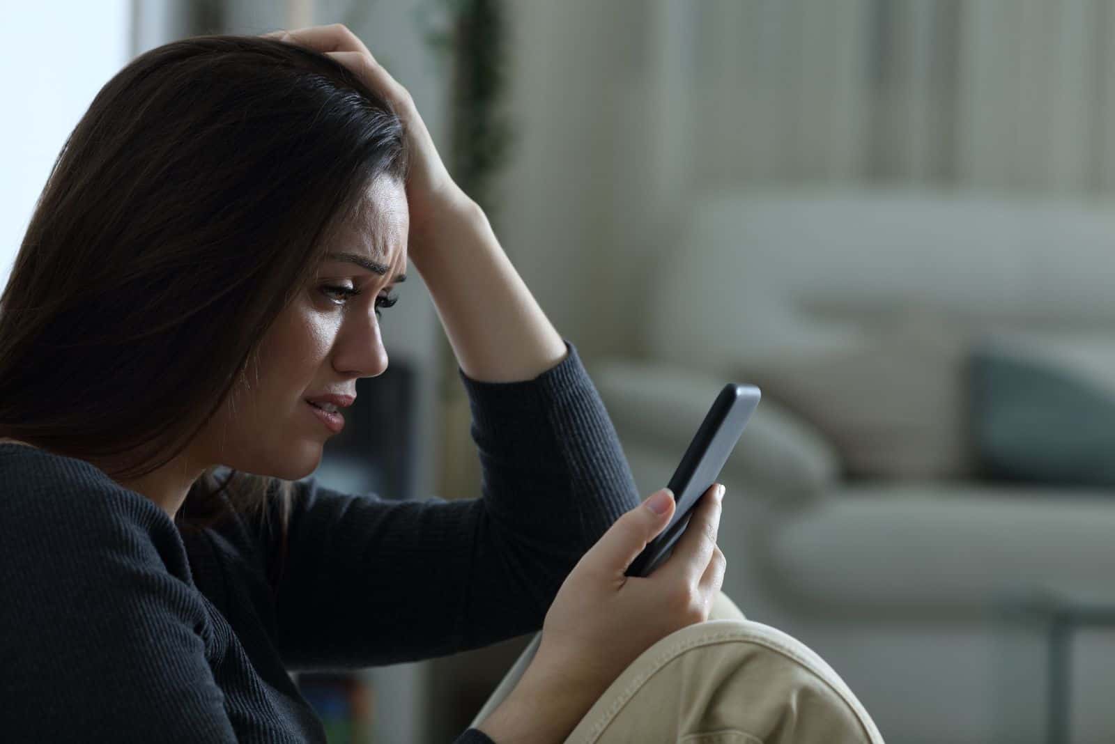 une fille aux longs cheveux noirs est assise sur le canapé en pleurant et tenant un téléphone portable à la main