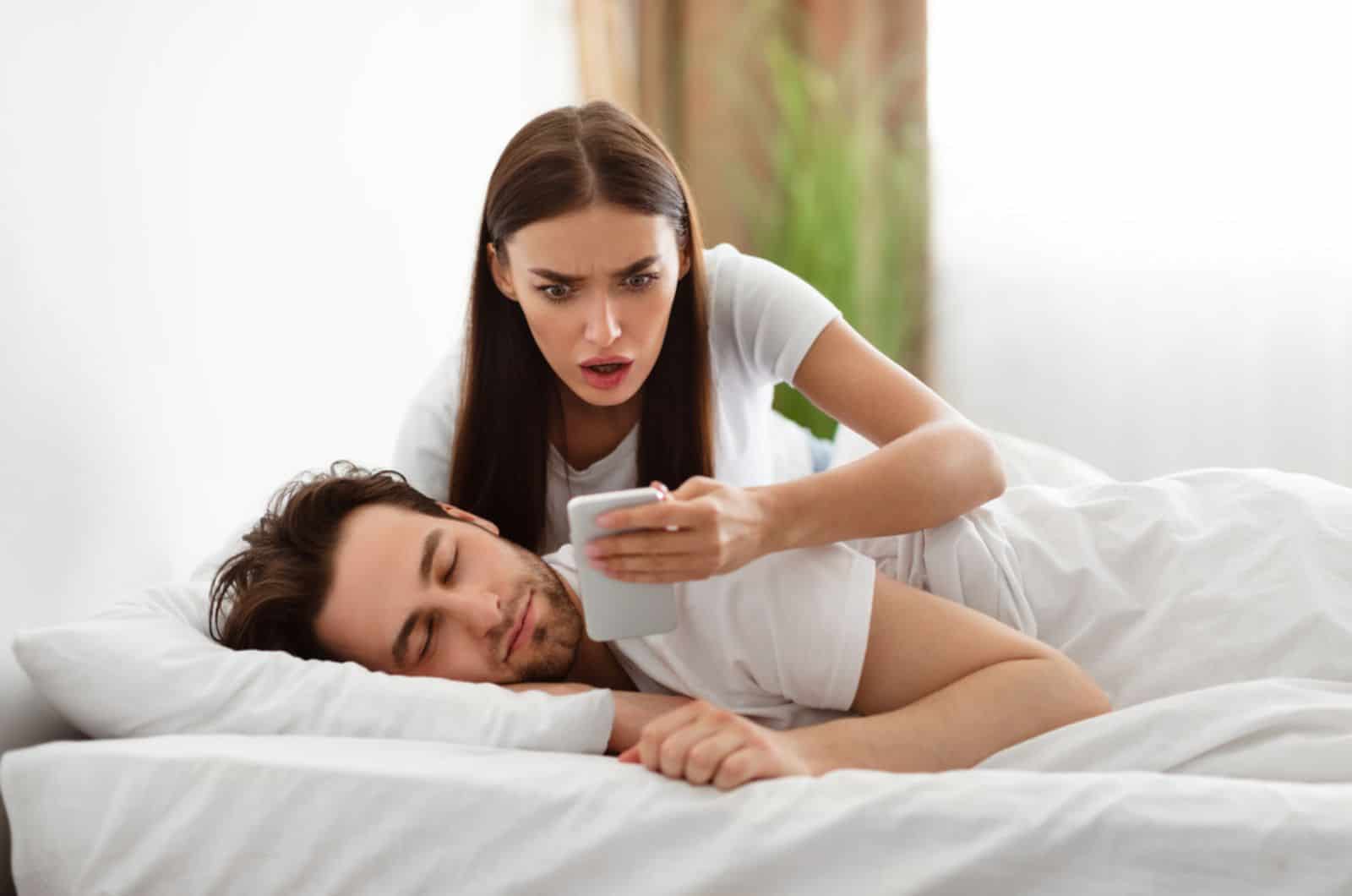Femme lisant un message de son amant au téléphone tout en trompant son mari en train de dormir