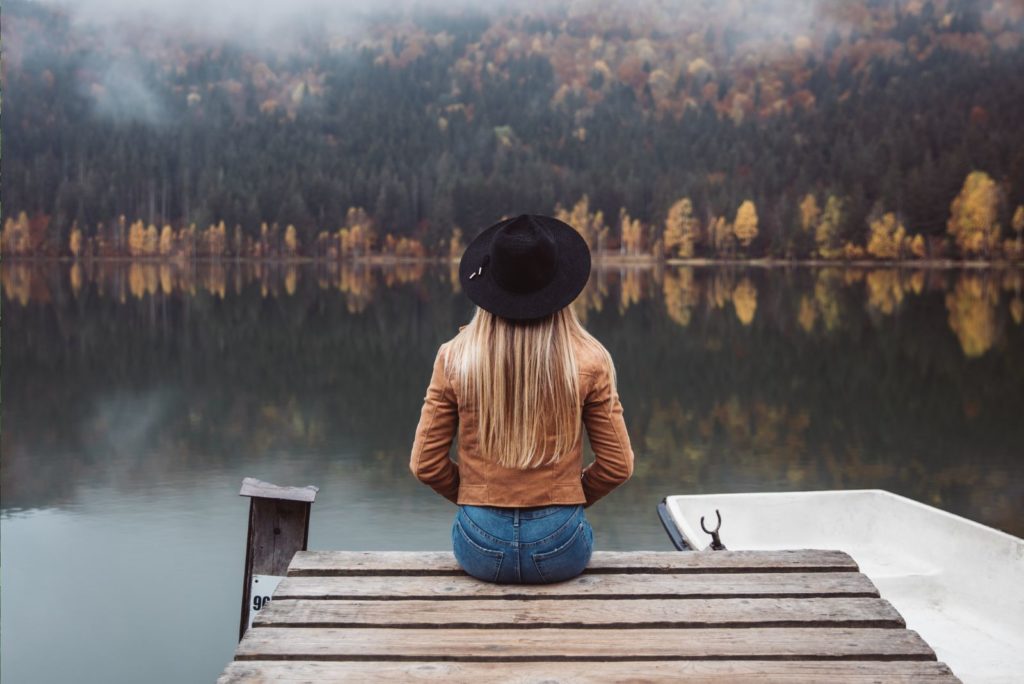 une fille aux longs cheveux blonds et un chapeau est assise sur la jetée